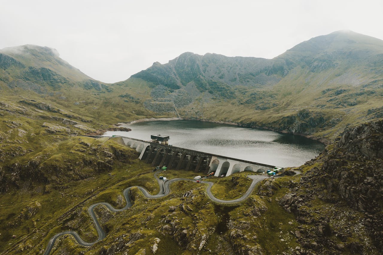 Scenic aerial shot of a curvy road and dam surrounded by mountains and a misty lake.